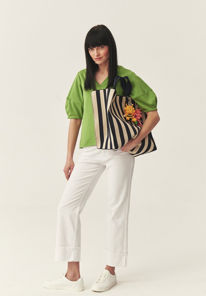 Femme aux cheveux noirs raides porte un haut vert, un pantalon blanc, des baskets blanches, et porte un sac fourre-tout rayé noir et beige avec des fleurs et un porte-clés.