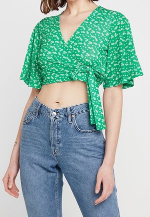Grünblümiges Crop-Top mit kurzen, flatternden Ärmeln und einem Wickeldesign, das an der Taille gebunden ist. Kombiniert mit hochgeschnittenen hellblauen Jeans.