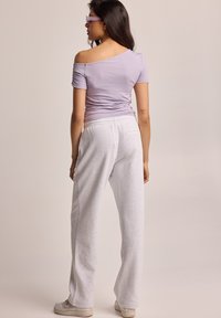 Haut violet clair à décolleté plongeant, ajusté avec des côtés froncés, associé à un pantalon de survêtement gris clair avec une poche arrière. Des baskets blanches complètent le look.