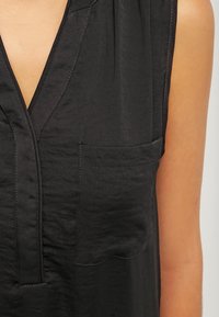Blouse sans manches noire avec un décolleté en V profond et une poche poitrine à l'avant, portée par une personne à la peau d'un ton marron clair.