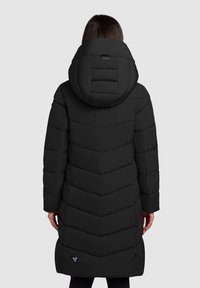 Vrouw in een lange zwarte gevoerde winterjas met een grote capuchon, gezien van achteren tegen een egale lichte achtergrond.