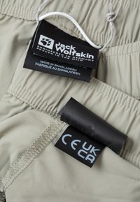 Shorts color khaki chiaro con vita elasticizzata. Presenta due etichette: logo 'Jack Wolfskin' e etichetta di certificazione 'UKCA'. Tessuto dalla consistenza liscia.