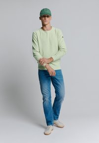 Lichtgroene langemouwen shirt van textuurstof, gedragen met lichtblauwe denimjeans en witte sneakers. Een groene pet maakt de outfit compleet.