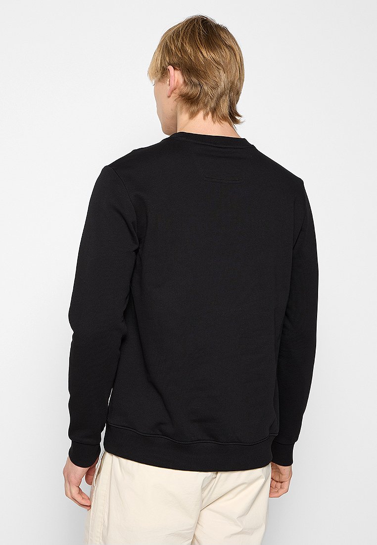 Persona bionda con indosso un semplice felpa nera a maniche lunghe e pantaloni beige chiaro, di spalle su uno sfondo bianco.