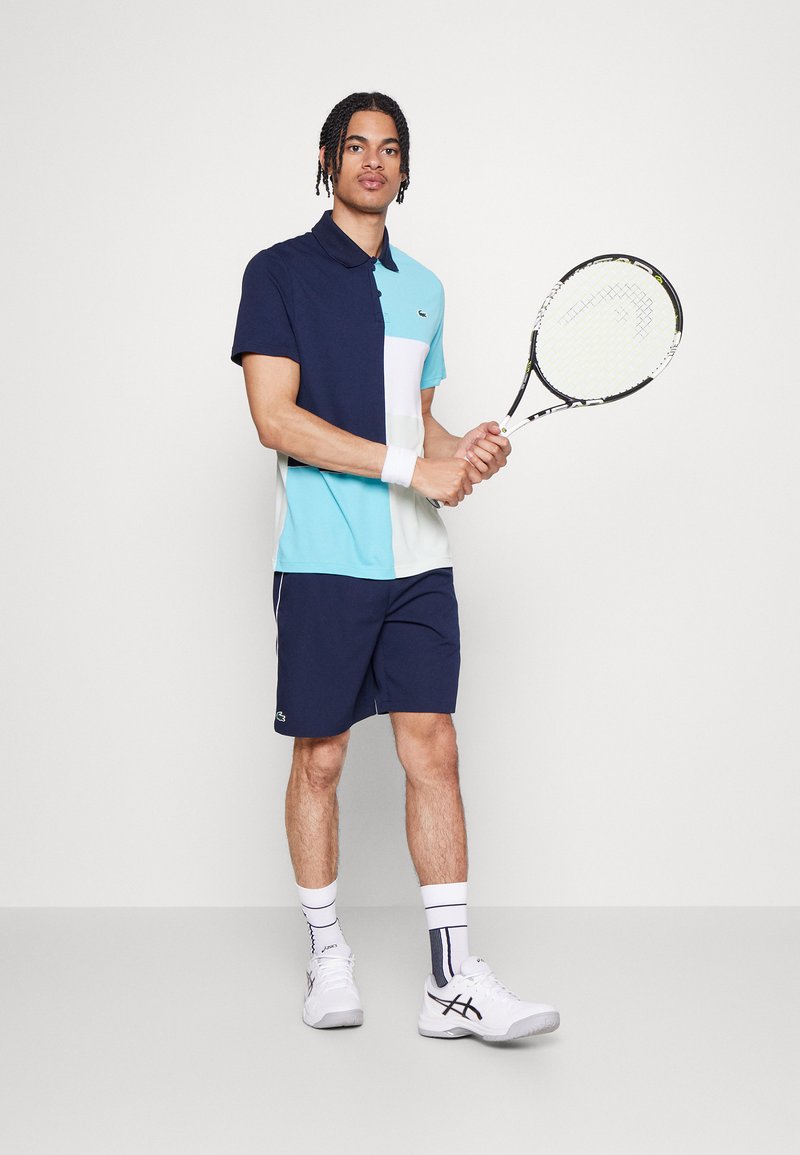 Tenue de tennis comprenant un polo coloré en bleu, marine et blanc, à manches courtes ; associé à un short marine et des baskets blanches.