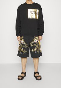 Svart sweatshirt med guldlogotyp, matchad med svarta shorts med färgglad blommiga broderier. Bärs med kraftiga svarta sandaler.