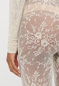 Σώμα από δαντέλα με μακριά μανίκια, λεπτό φλοράλ σχέδιο και κρέμ χρώμα. Το υλικό έχει διαφανή, υφασμάτινη εμφάνιση.