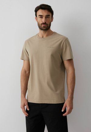 Mężczyzna o ciemnych włosach i brodzie, ubrany w beżowy t-shirt z krótkim rękawem i czarne spodnie, stojący na tle jednolitego jasnoszarego tła.