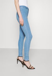 Jean skinny, em denim azul claro, com um design de cintura alta. Apresenta costuras mínimas e uma textura suave. Combinado com saltos altos pretos com tiras.