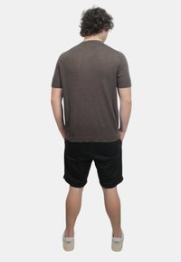 Camicia corta a maniche corte marrone con una vestibilità rilassata, indossata con pantaloni corti neri e scarpe chiare, che mostra un design semplice e informale.