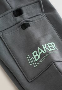 Grijze stoffen zak met een zwarte klittenbandsluiting. Bedrukte tekst "BAKER NINETEEN EIGHTY EIGHT" in mintgroen. Soepele textuur en strak ontwerp.