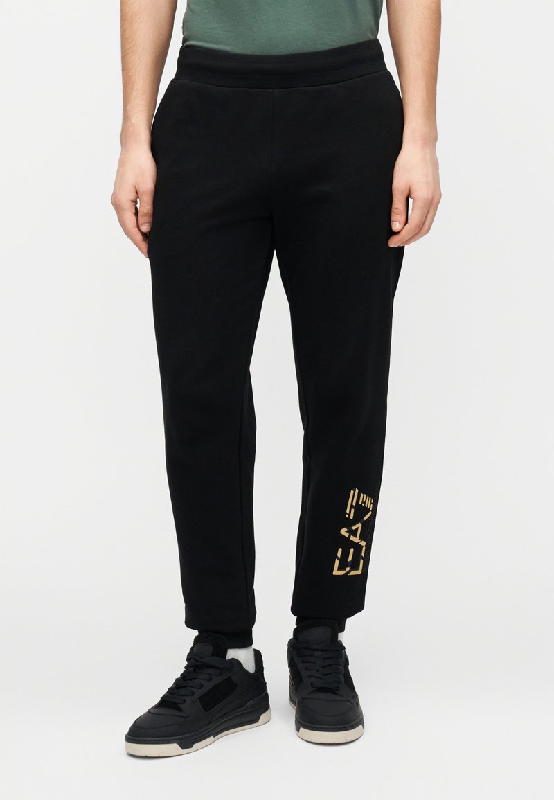 Čierne tepláky s rebrovaným pásom, zúženými nohavicami a zlatou grafickou potlačou na spodnej časti ľavej nohy. Kombinované s čiernymi teniskami.