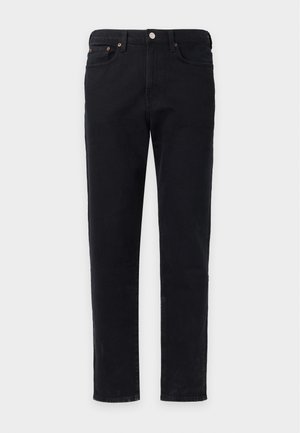 Jeune homme debout avec une main dans la poche, portant un t-shirt noir, un pantalon noir et des chaussures noires sans lacets, sur un fond clair uni.