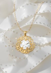 Guldkæde med en cirkul�ær vedhæng, der har en stor klar ædelsten, intrikat spiraldesign, hviler på hvid satin med små shiny accenter.