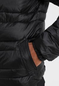 Czarna kurtka puchowa z pikowanym wzorem, elastycznymi mankietami i boczną kieszenią. Tekstura jest gładka i błyszcząca z widocznymi szwami.