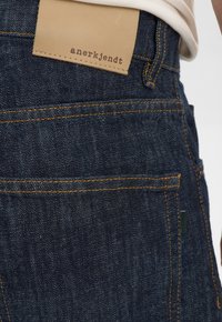 Jeans in denim scuro con cuciture arancioni a contrasto, etichetta in pelle con la scritta "anerkjendt" impressa e design slim fit.
