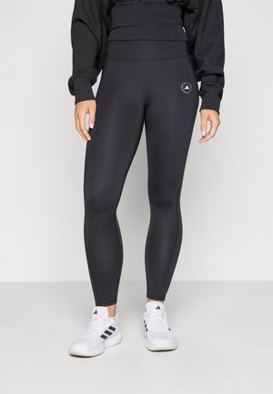 Black leggings made of stretchy material, featuring a high waistband and a small branded logo on the thigh. Worn with white athletic shoes.