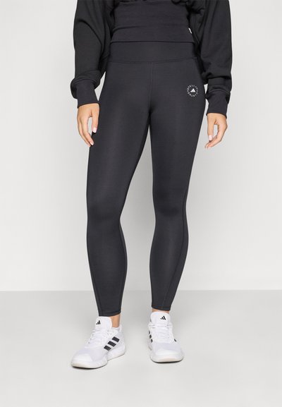 Leggings negros de material elástico, con una cinturilla alta y un pequeño logo de la marca en el muslo. Combinados con zapatillas deportivas blancas.