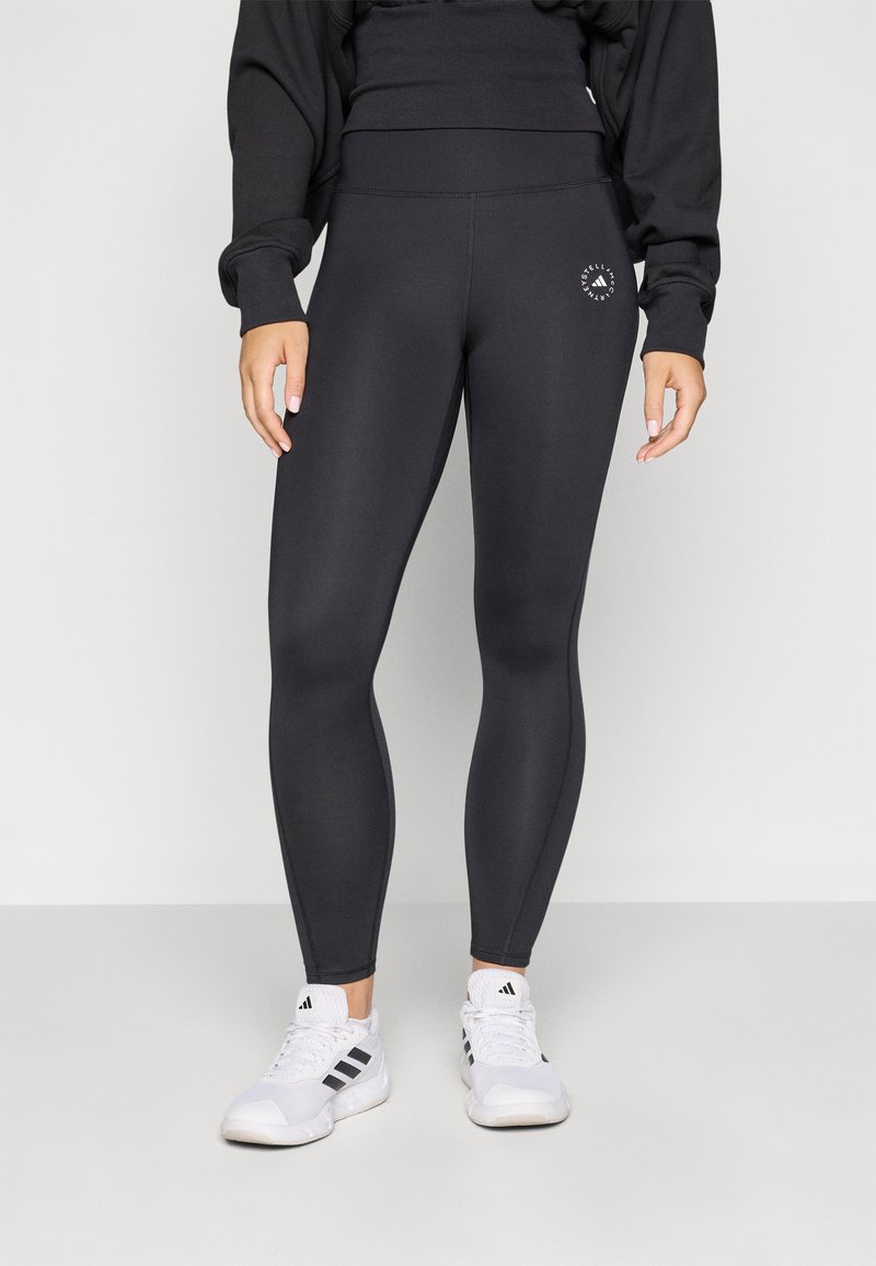 Leggings noirs en matériau extensible, avec une taille haute et un petit logo de marque sur la cuisse. Portés avec des chaussures de sport blanches.
