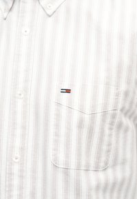 Λευκό πουκάμισο με κουμπιά, με λεπτές κάθετες γκρι ρίγες και μια μικρή τσέπη στο στήθος που διαθέτει λογότυπο σε κόκκινο, άσπρο και μπλε.