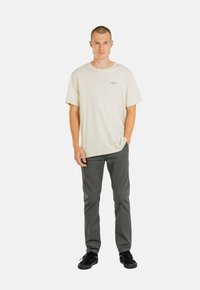 Camiseta de algodón color beige claro con cuello redondo y pequeño logo bordado en el pecho, combinada con pantalones oscuros grises y zapatillas negras.