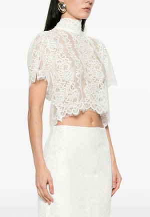 Donna che indossa un top corto trasparente in pizzo bianco con collo alto e maniche corte abbinato a una gonna in pizzo bianco a vita alta, grandi orecchini rotondi dorati visibili.