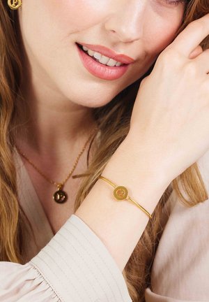 Gouden armband met een ronde hanger met een verhoogde letter "P." Gladde metalen afwerking, gedragen om een pols naast een bijpassende gouden hangerketting.