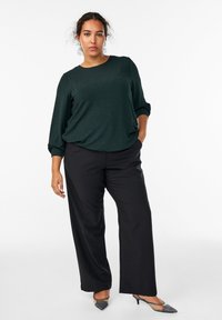 Ciemnozielona bluzka z długim rękawem o luźnym kroju, w połączeniu z czarnymi spodniami o szerokich nogawkach i szarymi butami na obcasie. Gładka tekstura, minimalne detale.