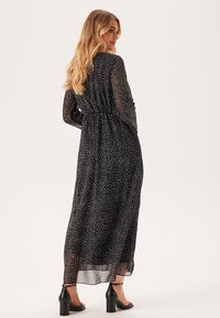 Robe maxi noire avec de longues manches transparentes, taille élastique et motif à pois blancs. Longueur en dessous de la cheville, associée à des chaussures noires à talons.