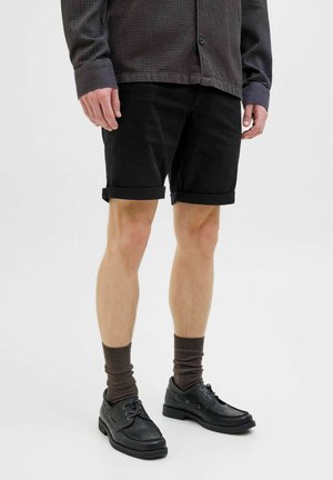 Schwarze Shorts mit umgeschlagenen Säumen, kombiniert mit braunen gerippten Socken und schwarzen Schnürschuhen mit subtilen Hardwaredetails. Texturiertes Hemd sichtbar.