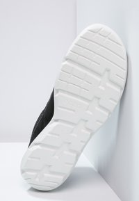 Černá teniska s texturovaným černým svrškem a bílou gumovou podrážkou se vzorovaným dezénem pro lepší přilnavost. Logo značky je embosováno na boku.