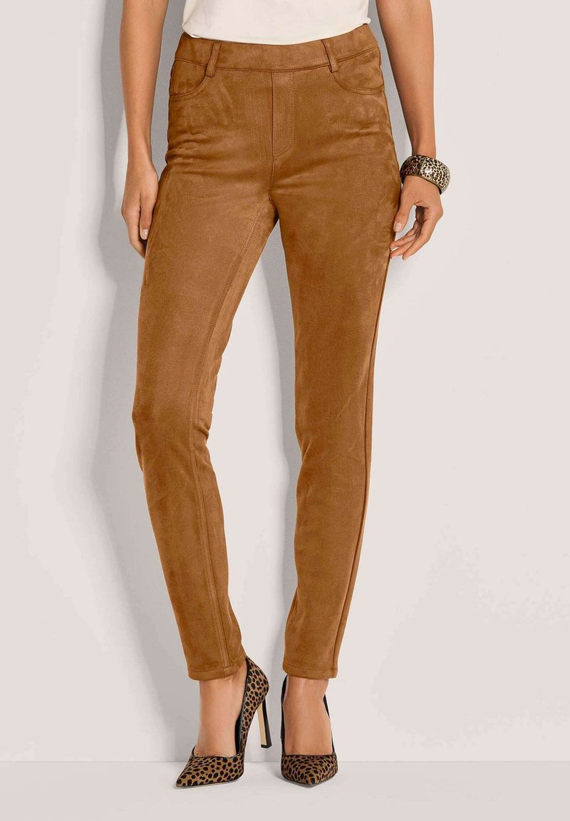 Vrouw in bruine suède skinny broek, met lederen hoge hakken in luipaardprint en bijpassende lederen armband in luipaardprint, staand tegen een lichte achtergrond.