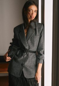 Blazer gris texturizado con cintura entallada y cinturón negro, combinado con un bolso negro con diseño estampado.