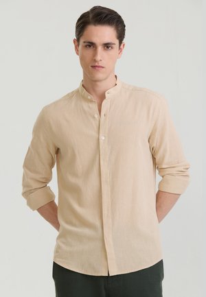 Młody mężczyzna o ciemnych włosach, ubrany w beżową koszulę z kołnierzykiem typu mandarynka i podwiniętymi rękawami, stoi na jasnym jednolitym tle.