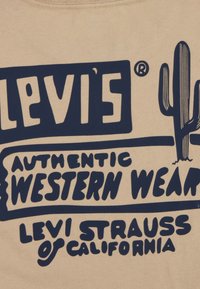 Camiseta beige con estampado en azul oscuro que presenta "Levi's", "Auténtica Ropa Occidental" y un diseño de cactus; fabricada en suave algodón.