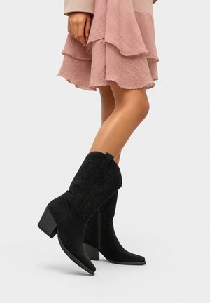 Woman wearing black suede mid-calf cowboy boots with embroidered details paired with a pink layered skirt and beige jacket.
