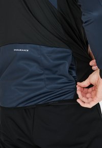 Svart och marinblå träningslinne med en texturerad yta, som har en sidficka och "ENDURANCE"-logotyp, designad för synlighet och komfort vid aktiv användning.