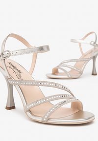 Silberne Sandalen mit mehreren überkreuzten Riemen, metallischem Finish, quadratischen Verzierungen, offenem Zehenbereich und schmalem Stiletto-Absatz.