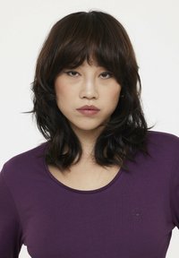 Haut à col rond violet à manches longues en tissu doux, avec un petit logo circulaire en bas à gauche. Texture lisse, design ajusté.