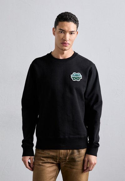Sweatshirt noir avec un logo brodé vert au niveau de la poitrine, à col rond et à manches longues, associé à un pantalon marron.