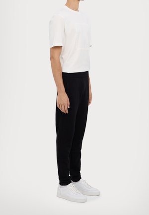 Bílý bavlněný tričko s krátkým rukávem a grafickým designem; v kombinaci s černými tepláky a bílými teniskami. Jednoduchý, neformální outfit.