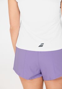 Vista posteriore di una persona che indossa una maglietta senza maniche bianca con un piccolo logo nero e pantaloni sportivi leggeri viola contro uno sfondo uniforme.