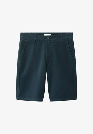 Mladý muž oblečený v béžovej bunde cez čierno-biele pásikavé tričko a tmavomodré šortky, stojaci pred bielym pozadím.