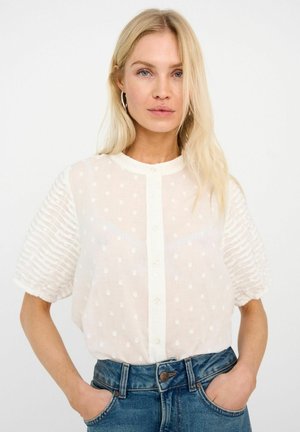 Weiße Bluse mit weißen Punkten, kurzen, gepufften, gestreiften Ärmeln und einer geknöpften Vorderseite. Leichter, halbtransparenten Stoff. Getragen mit hoch taillierter blauer Jeans.