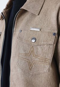 Chaqueta de denim marrón con acabado texturizado, que presenta un bolsillo en el pecho con un patrón de costura en forma de estrella y una etiqueta de logo plateada. Cierre con cremallera.