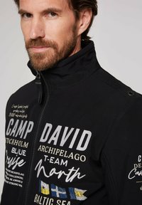 Sort lynlåsjakke med hvid og beige tekst, høj krave, broderede detaljer og farverige flagdesigns på den nederste front.