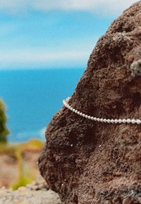 Weiße Perlenarmband ruht auf einem strukturierten dunklen Stein, mit einem verschwommenen blauen Ozeanhintergrund im Blick. Der Gesamtschwerpunkt liegt auf dem Armband und dem Stein.