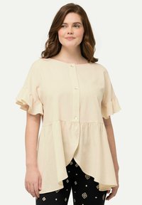 Blusa beige chiaro con scollo rotondo, chiusura a bottoni sul davanti e maniche corte voluminose. Il tessuto ha una consistenza morbida; orlo asimmetrico.