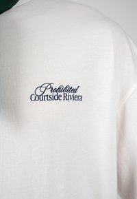 Άσπρο ύφασμα με μπλε σκούρο κείμενο «Prohibited Courtside Riviera» τυπωμένο στο άνω μέρος της πλάτης ενός ενδύματος.