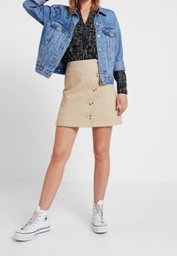 Giacca di jeans, blusa nera a fantasia, gonna beige con dettagli a bottoni e sneaker bianche alte. Outfit casual e stratificato.